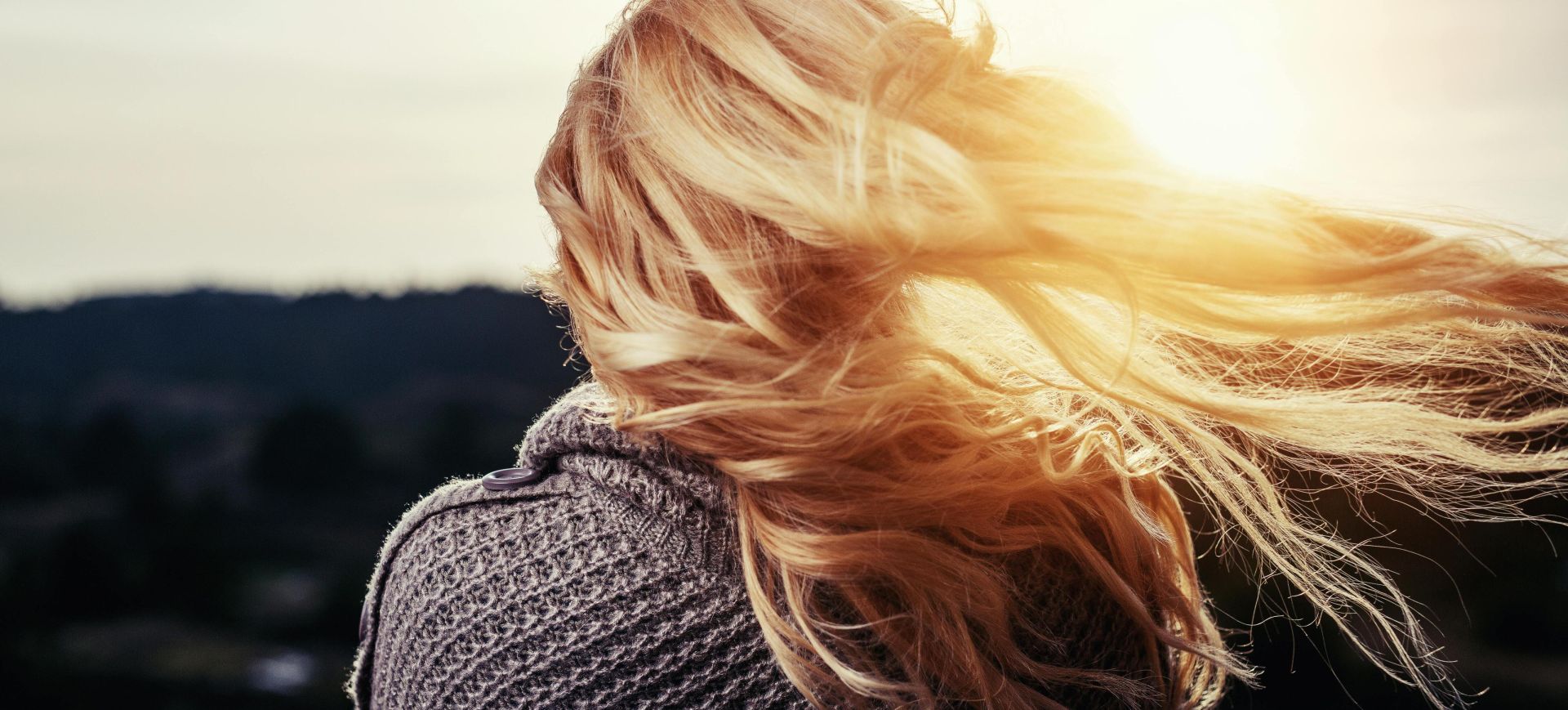 Back view of a woman with blonde hair blowing in the wind during sunset outdoors.